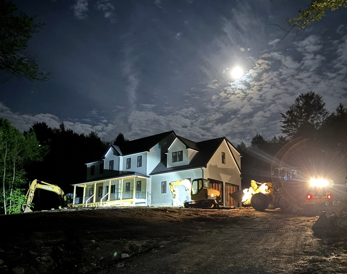 New England Developments home under construction at night, lit by equipment headlights with a full moon overhead.