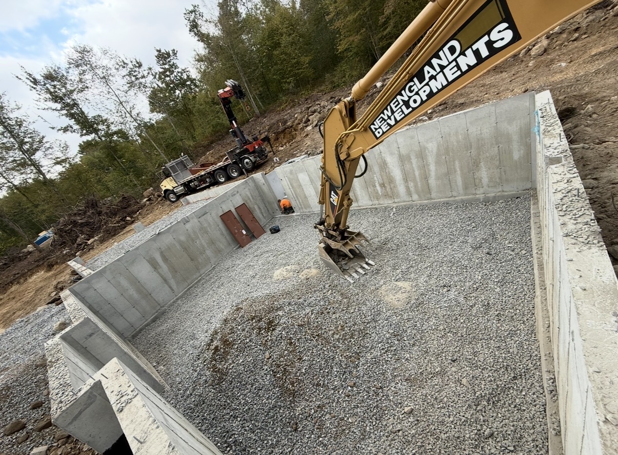 A New England Developments excavator working inside a poured concrete foundation.